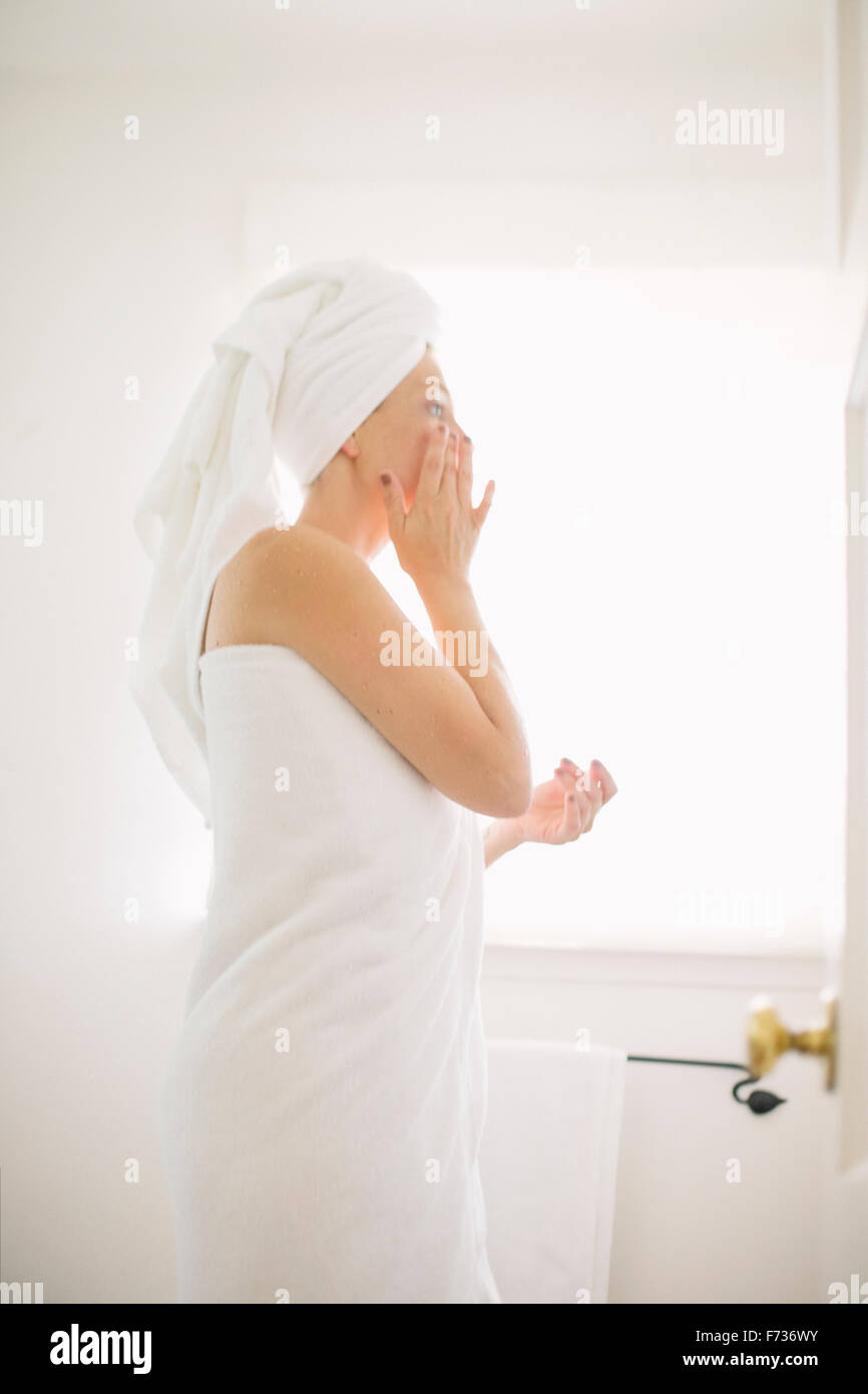 Donna avvolta in un asciugamano bianco in piedi in un bagno, l'applicazione di crema per il viso. Foto Stock