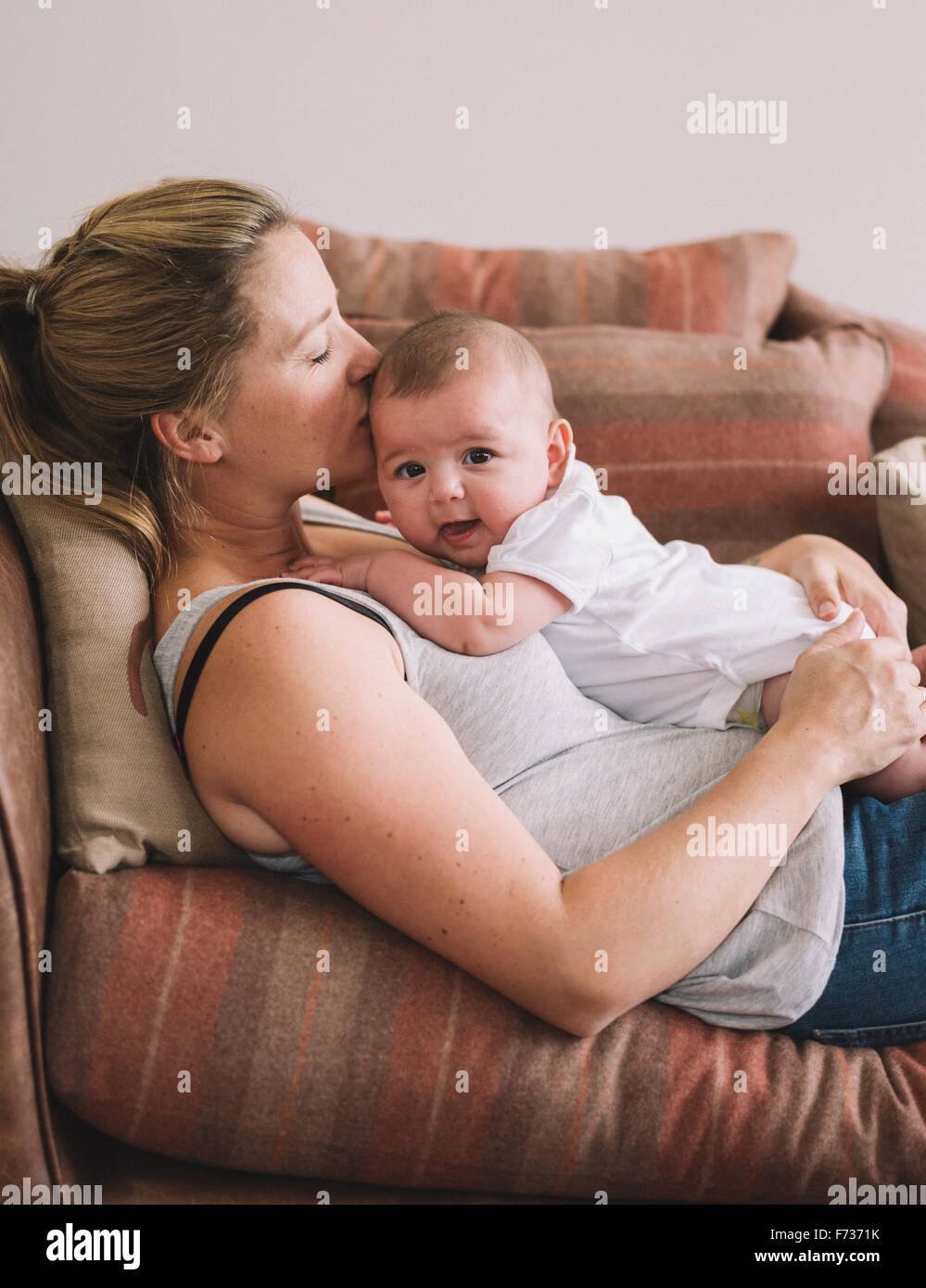 Una donna sdraiata su un divano a giocare con una bambina, baciando la sua testa. Foto Stock