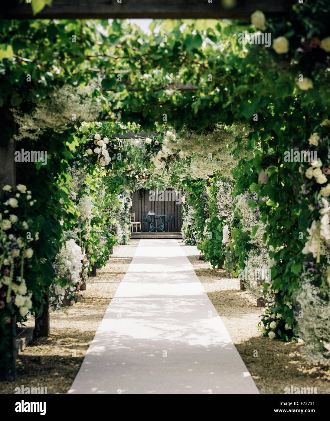 Un giardino percorso attraverso un arco che conduce a un'alcova con tavolo e sedie, con piante rampicanti e rose bianche. Foto Stock