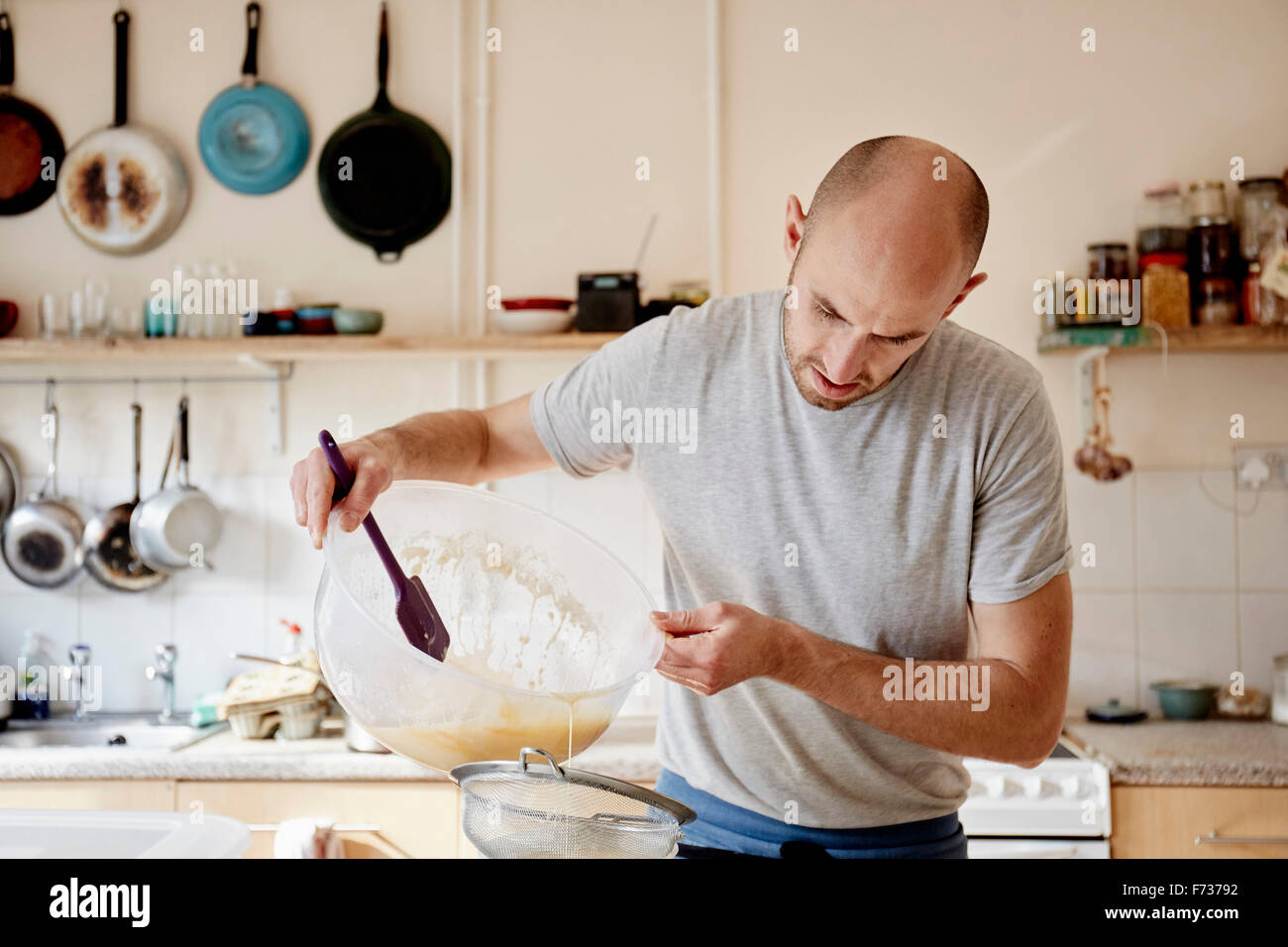 Un baker lavora in una cucina, versando un liquido in un recipiente attraverso un setaccio, rendendo l'impasto. Foto Stock