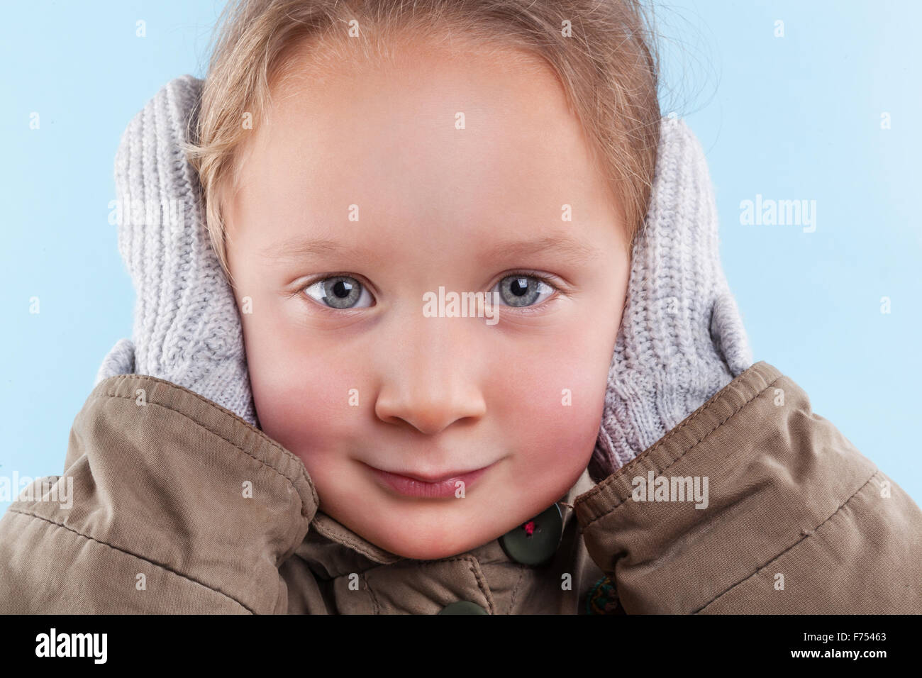 Piccola ragazza carina che copre le sue orecchie con le mani in guanti protettivi contro la luce sfondo blu. Stagione invernale. Foto Stock