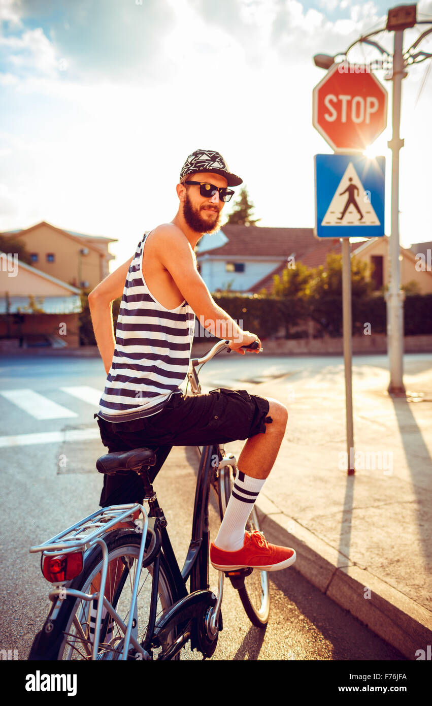 Giovane uomo elegante in occhiali da sole di una bicicletta su una strada di città alla luce del tramonto Foto Stock
