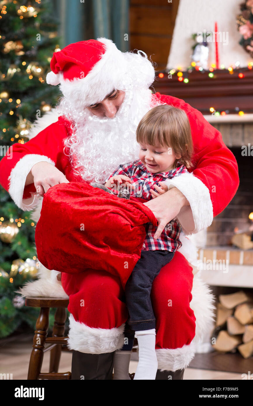 Babbo Natale dando un presente dal sacco a kid boy vicino al camino e albero di Natale a casa Foto Stock