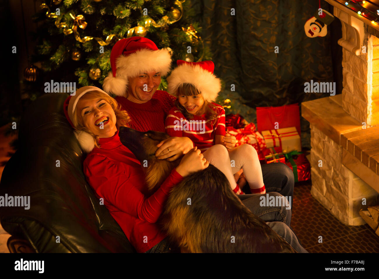 La famiglia felice in cappelli rossi con cute cane seduti ad albero di Natale vicino al camino Foto Stock