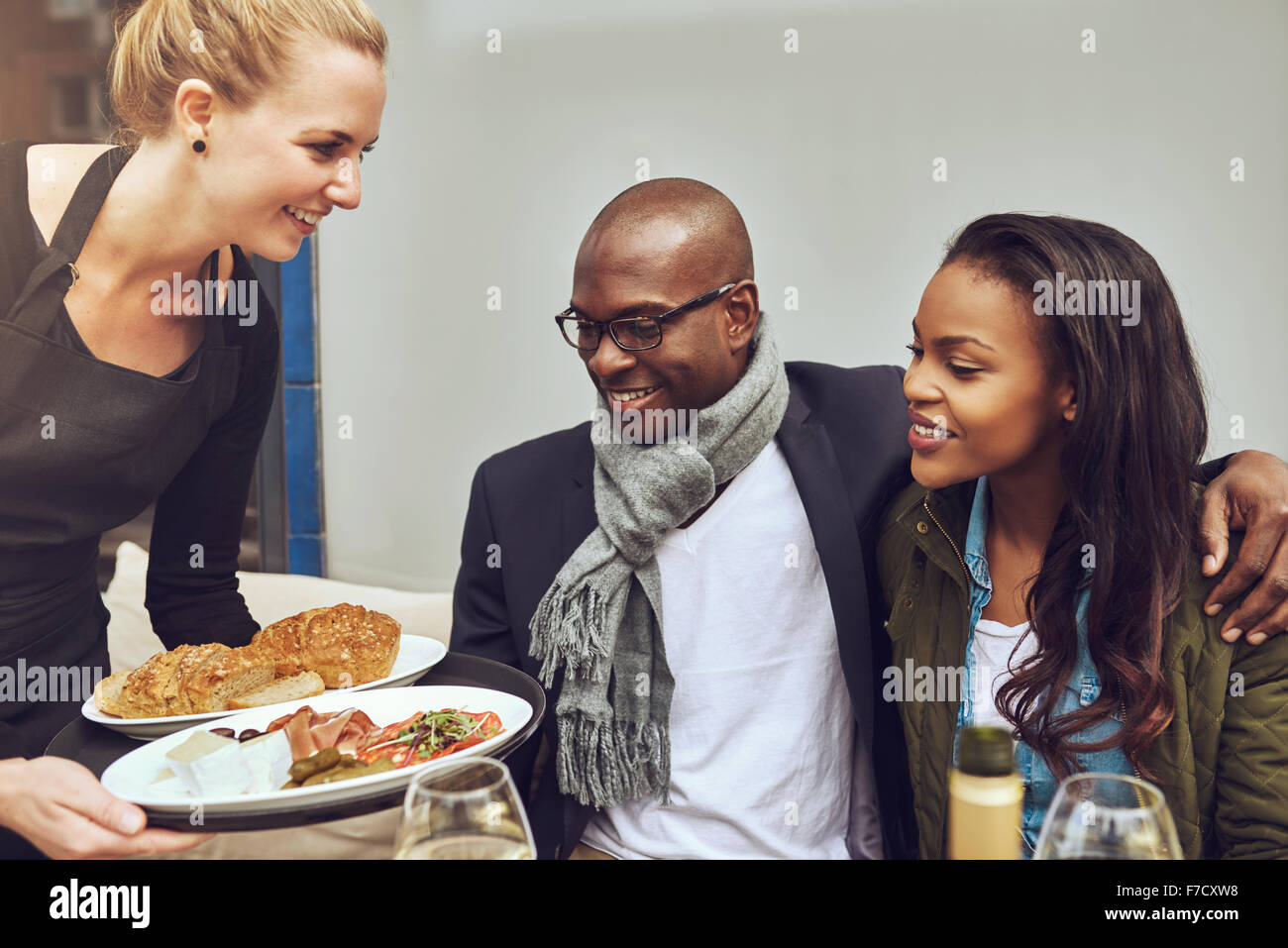 Attraente giovane americano africano giovane seduto a braccetto a una tavola di ristorante viene servita la cena da un giovane sorridente Caucasi Foto Stock