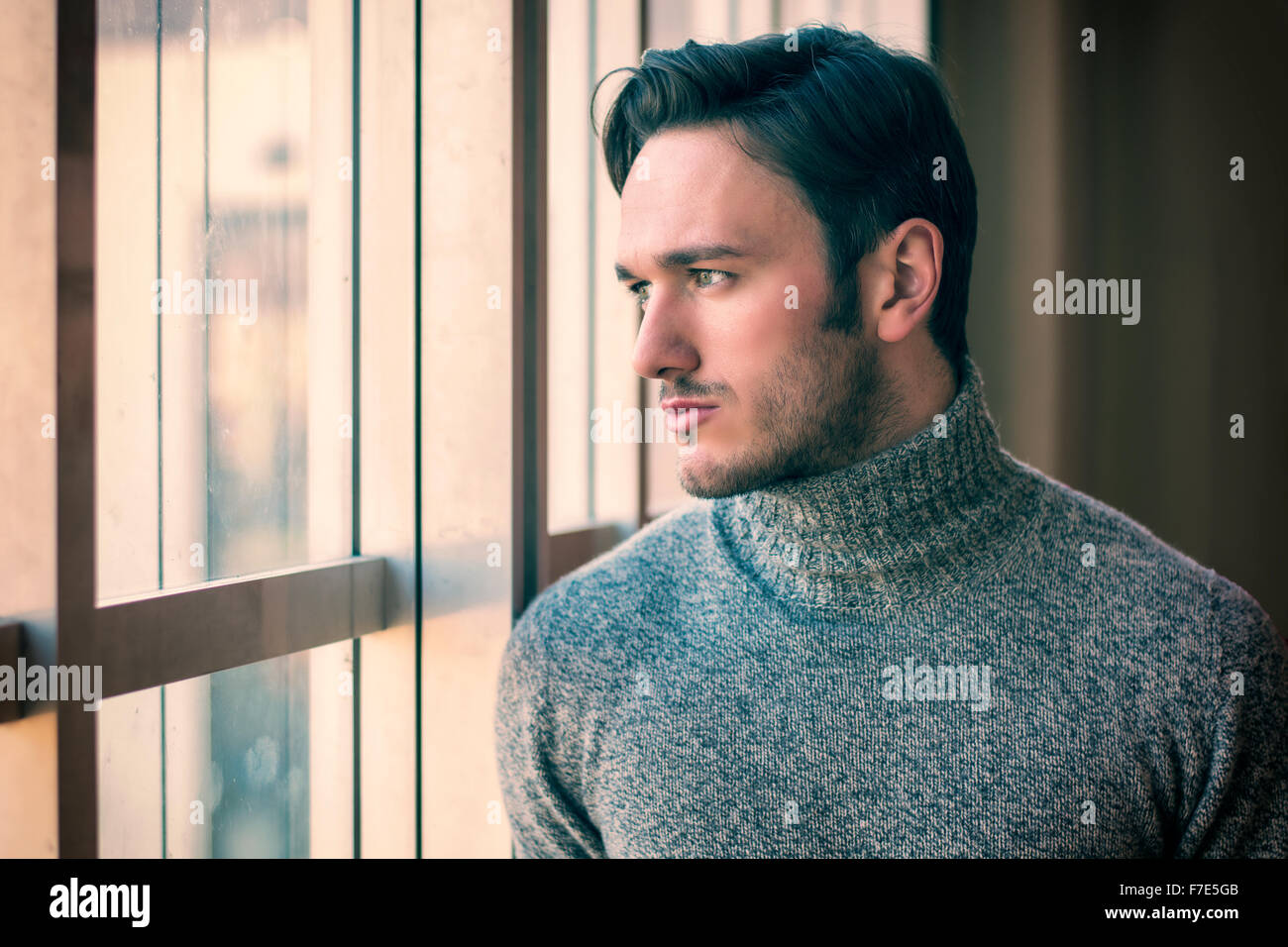 Bello grave uomo in piedi all'interno di edificio moderno accanto alla finestra grande, indossare un maglione di lana, guardando fuori Foto Stock