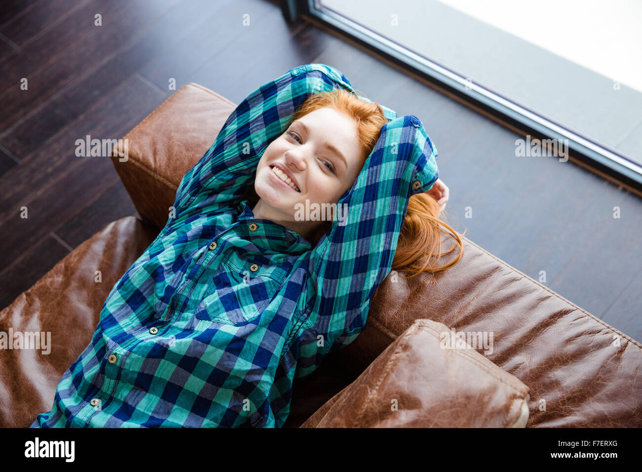 Rilassato positivo redhead girl in blue plaid shirt appoggiato sulla pelle marrone lettino Foto Stock