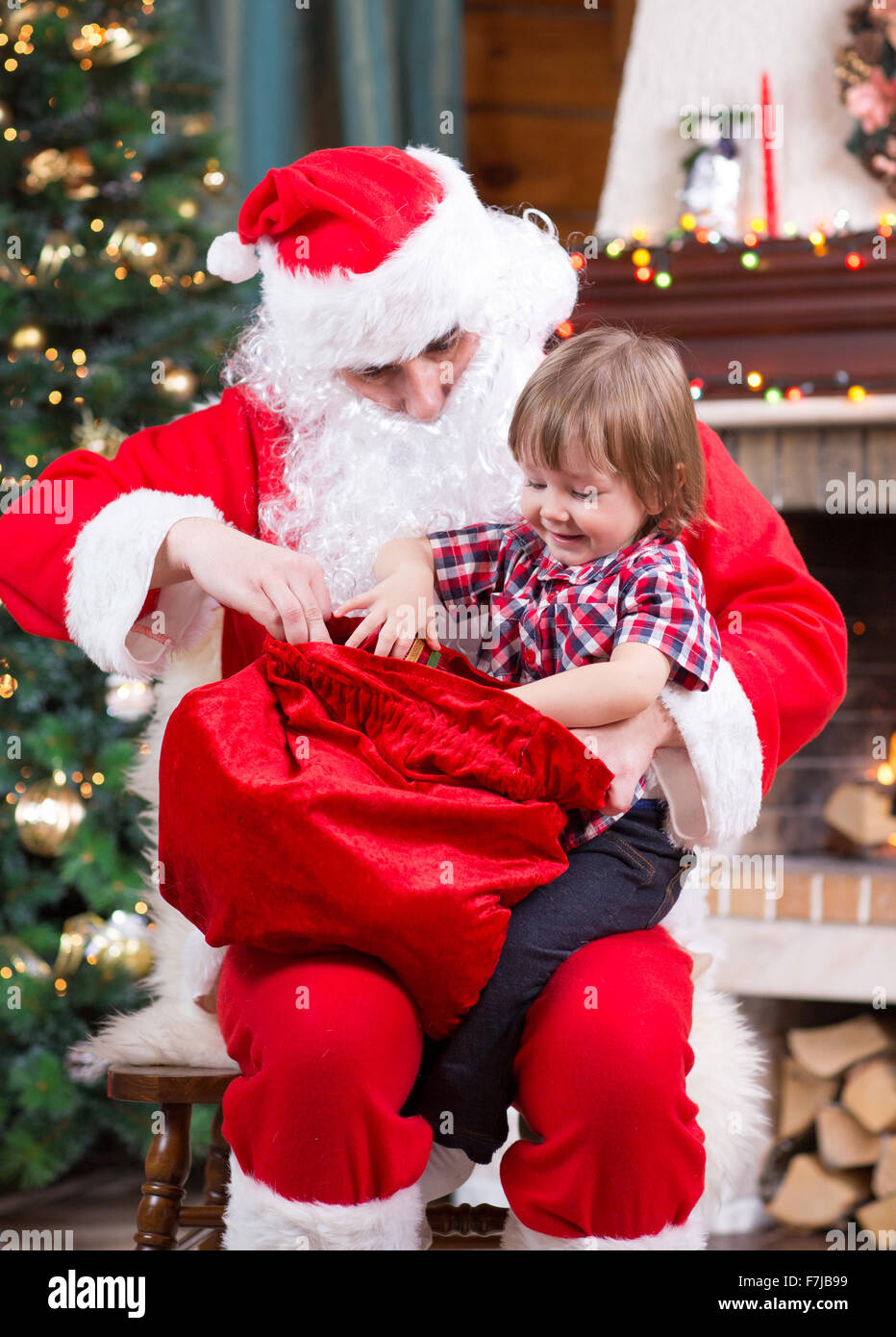 Santa Claus tira un presente fuori sacco al bambino vicino al camino e albero di Natale a casa Foto Stock