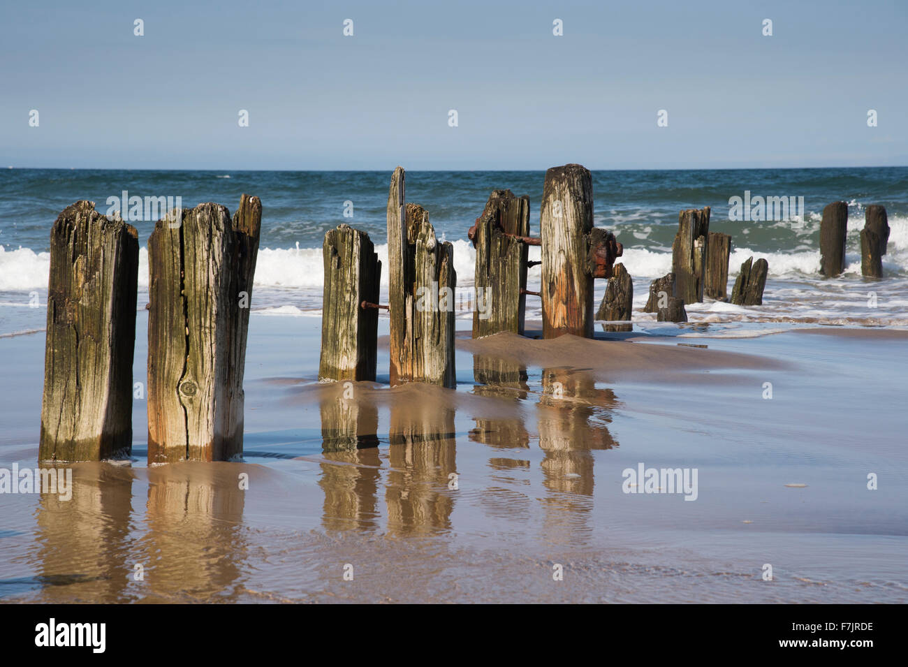 Summer sun & blue sky. Mare onde del giro la battigia mentre vecchi pennelli di legno sono riflessi nell'acqua. Sandsend spiaggia costa dello Yorkshire, Inghilterra, Regno Unito. Foto Stock