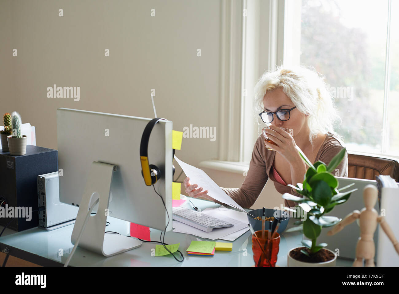 Giovane donna bere il tè e la lettura di documenti cartacei alla scrivania in ufficio in casa Foto Stock