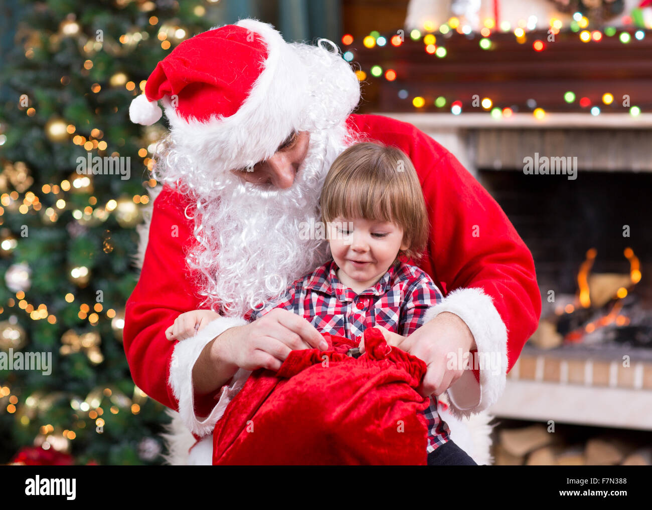 Babbo Natale dando un presente dal sacco al bambino ragazzo vicino al camino e albero di Natale a casa. Foto Stock