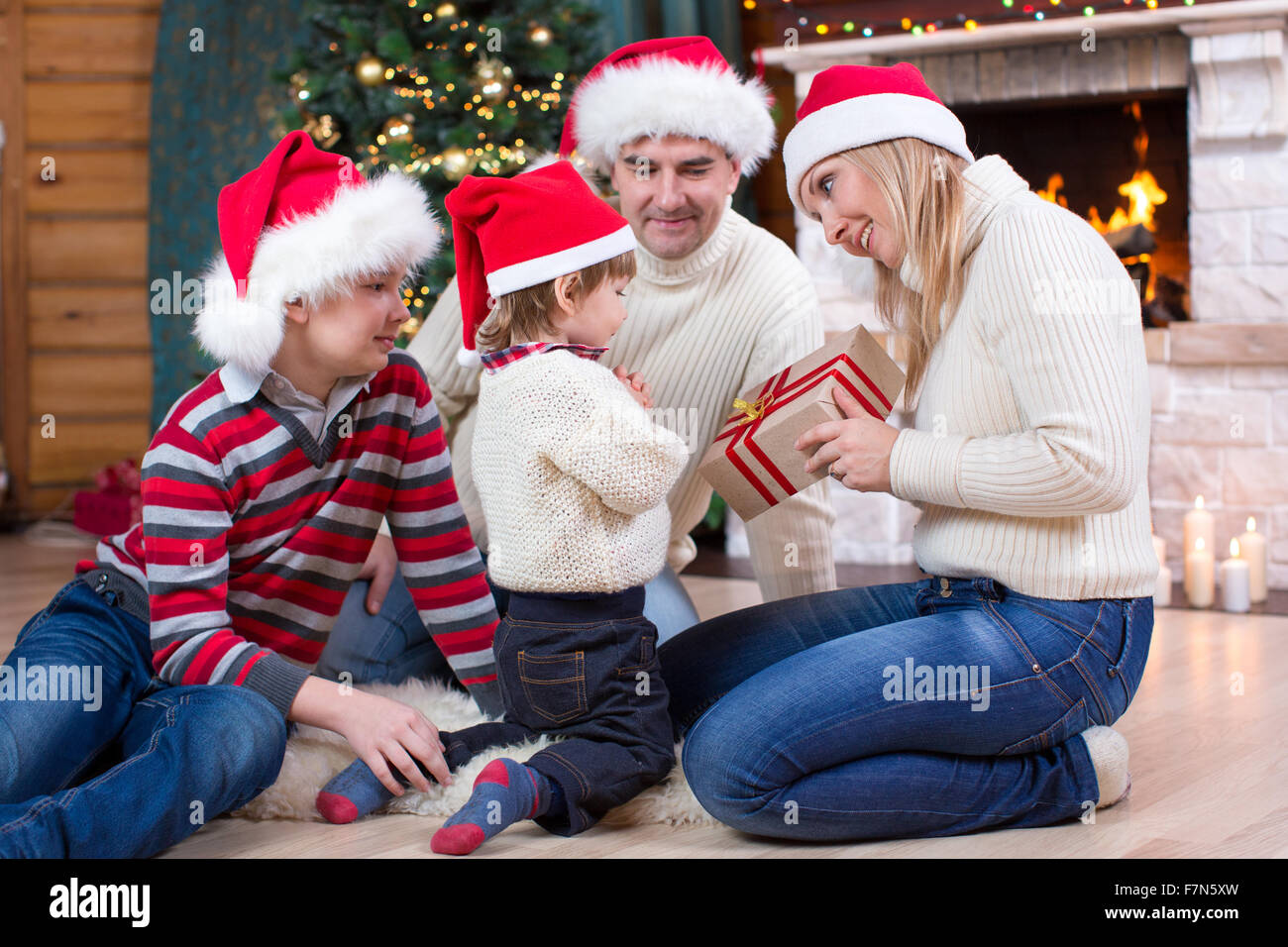 La famiglia felice in cappelli rossi con dono seduti ad albero di Natale vicino al camino Foto Stock