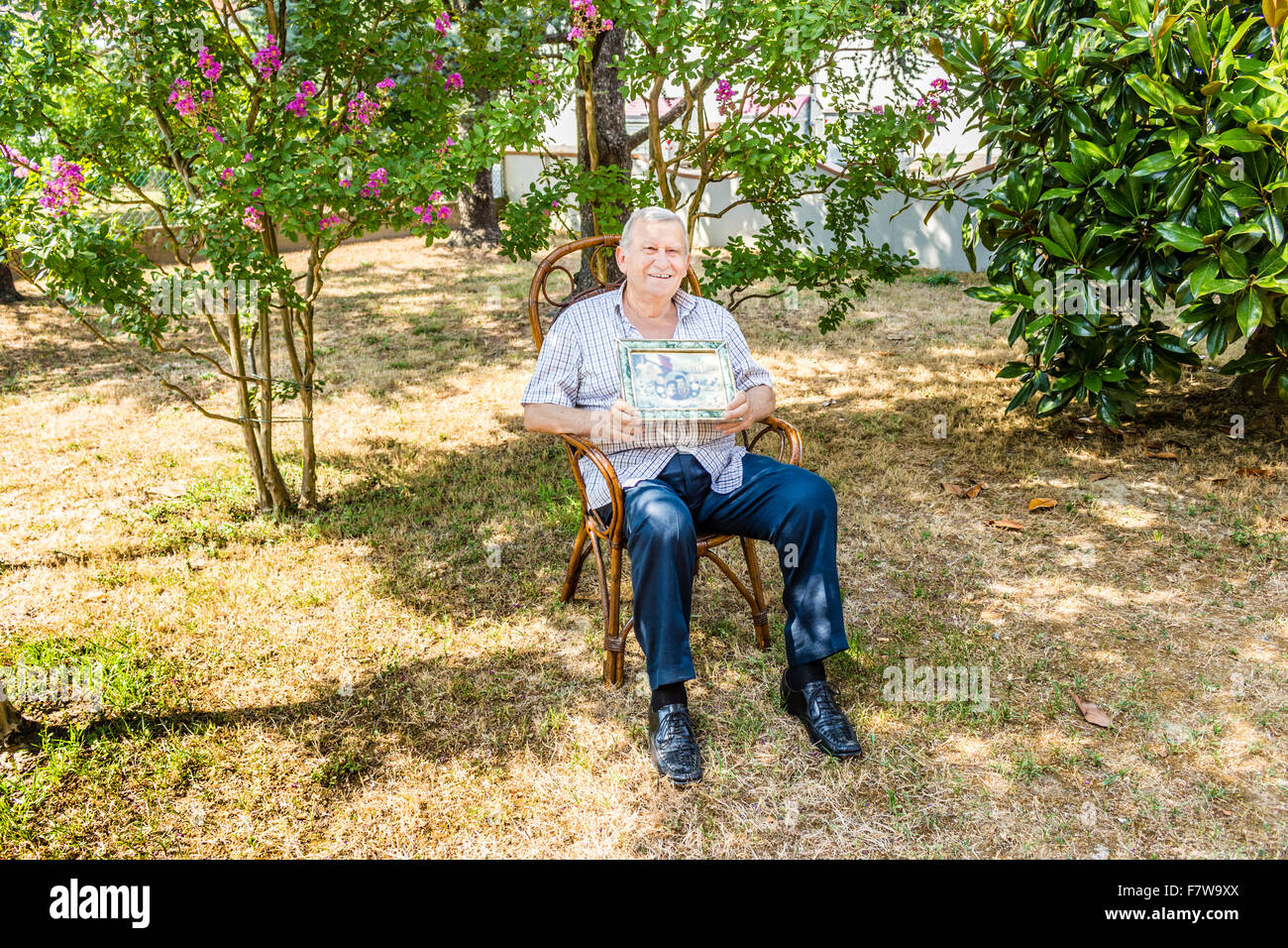 Anziani maschi octogenarian sorridente e mostrando il vecchio vintage foto in una cornice mentre è seduto sulla sedia di vimini in giardino Foto Stock