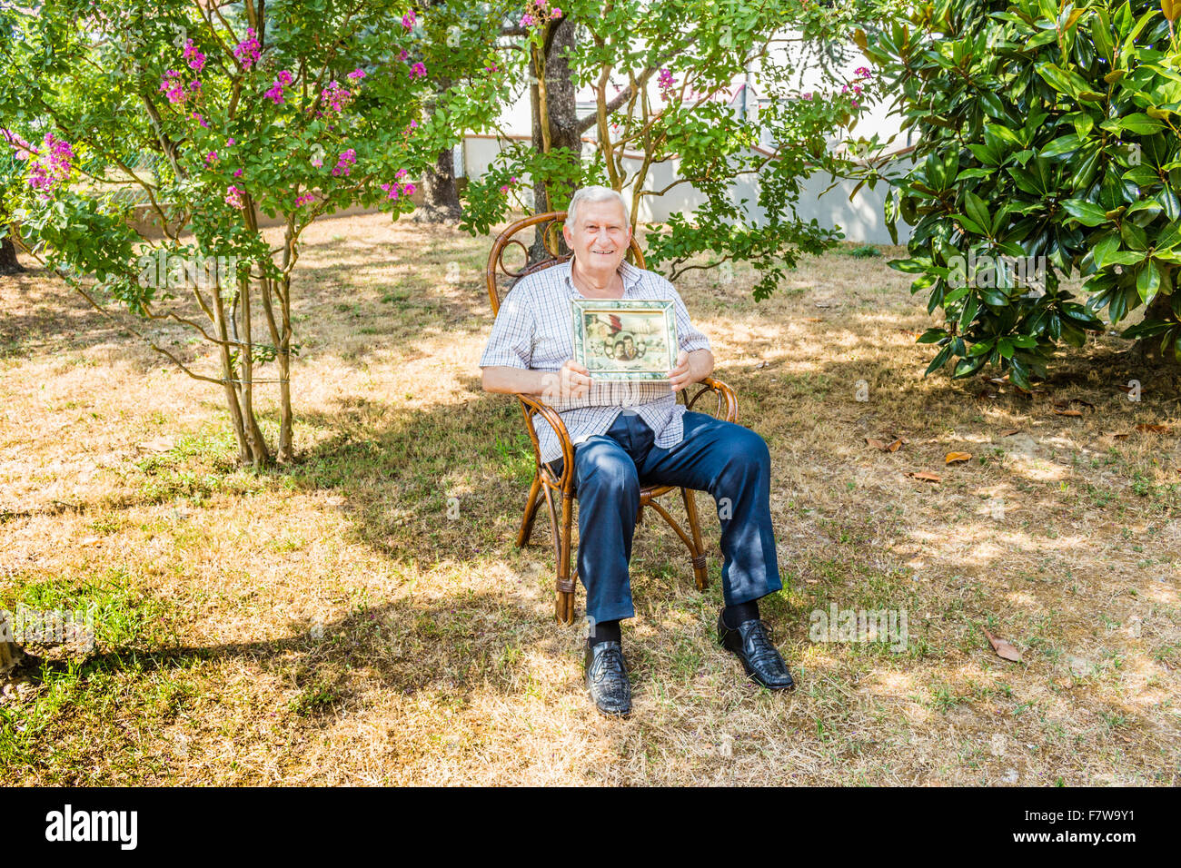 Anziani maschi octogenarian sorridente e mostrando il vecchio vintage foto in una cornice mentre è seduto sulla sedia di vimini in giardino Foto Stock