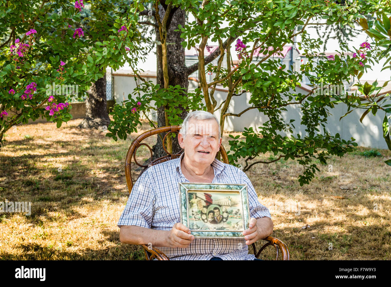 Anziani maschi octogenarian sorridente e mostrando il vecchio vintage foto in una cornice mentre è seduto sulla sedia di vimini in giardino Foto Stock