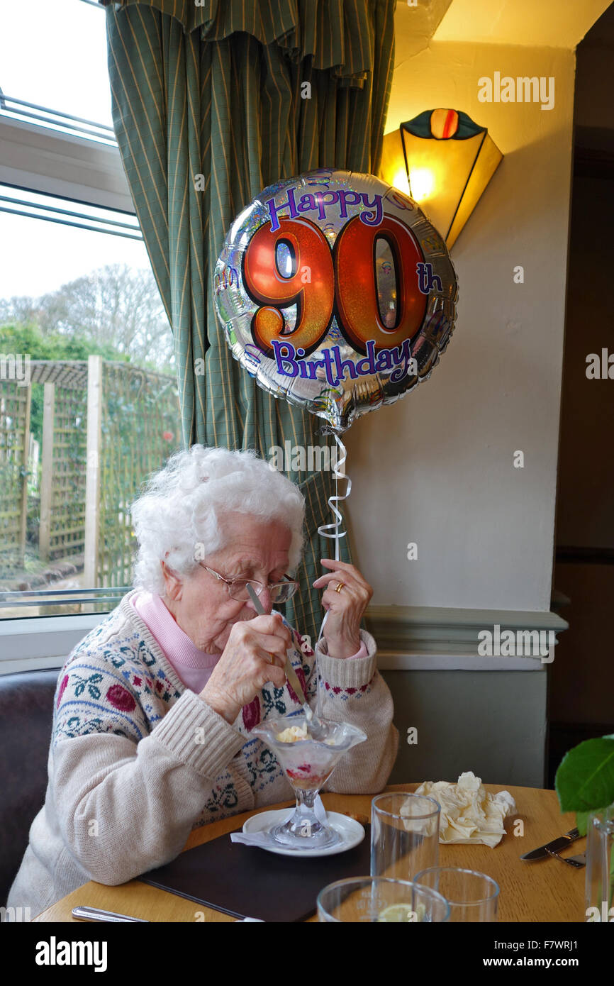 Una donna anziana che celebra il suo novantesimo compleanno di mangiare un gelato Gelato e tenendo un palloncino con felice il novantesimo compleanno su di esso, England, Regno Unito Foto Stock