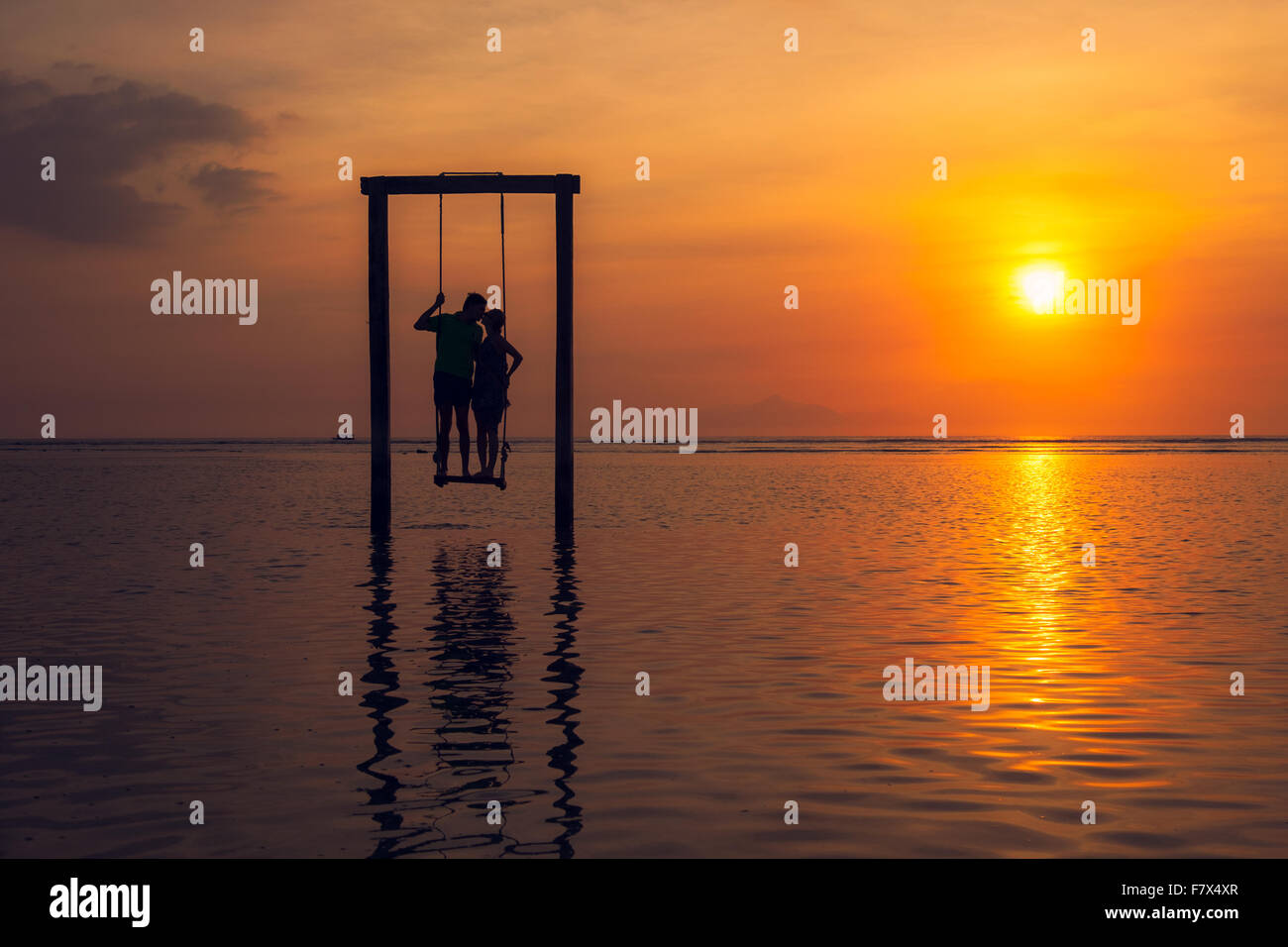 Silhouette di un uomo e di una donna baciare, in piedi su un altalena in mare al tramonto, Indonesia Foto Stock