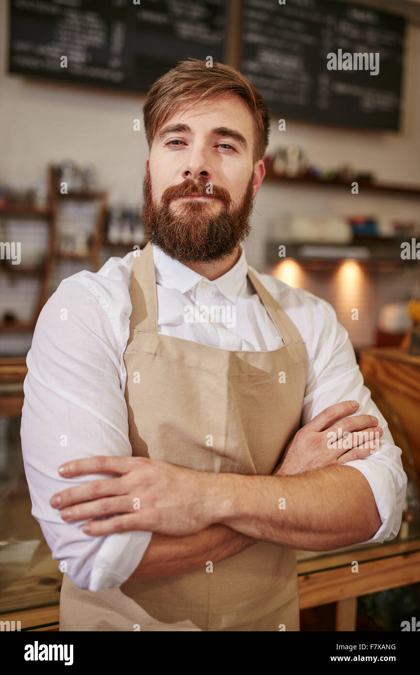 Ritratto di giovane uomo in piedi in un coffee shop con le braccia incrociate. Uomo caucasico con la barba in piedi in una caffetteria guardando ca Foto Stock