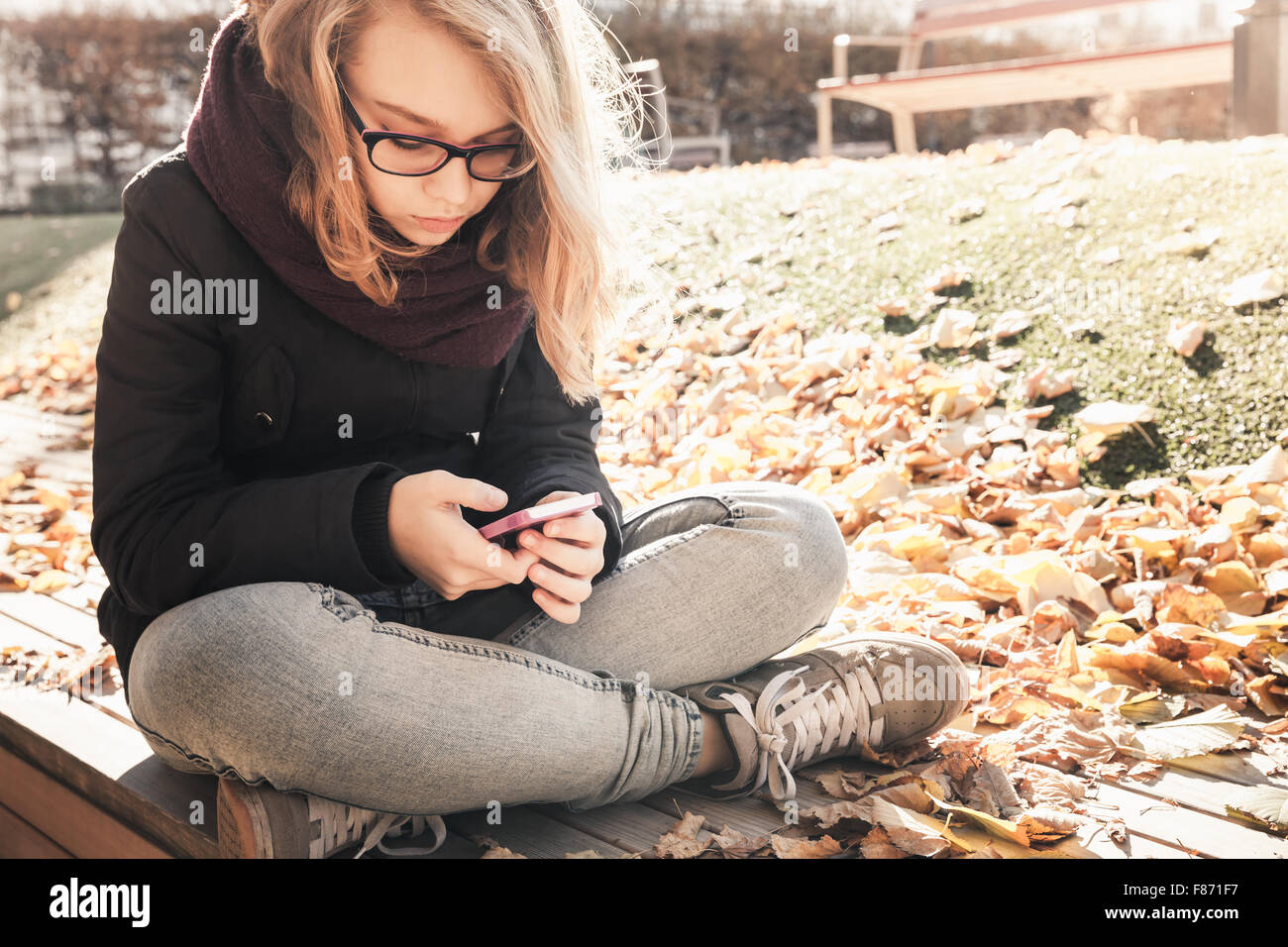 Bionda caucasica ragazza adolescente in jeans e giacca nera seduta in legno su una panchina nel parco e utilizza lo smartphone, outdoor autunno Foto Stock