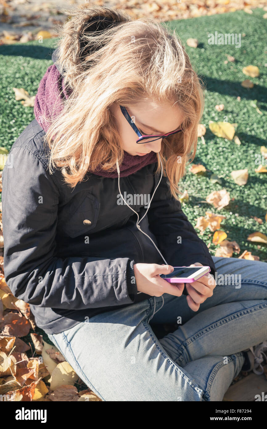 Bionda caucasica ragazza adolescente in jeans e giacca nera seduto in posizione di parcheggio e utilizza lo smartphone, outdoor autunno ritratto, vintage st Foto Stock