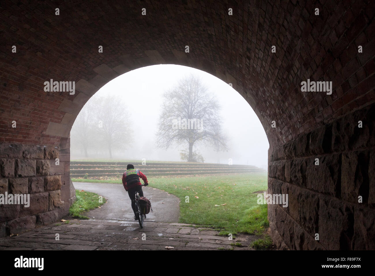 Ciclista escursioni in bicicletta su un percorso di ciclo ed emergenti da sotto l'arco di un ponte su un nebbioso giorno in autunno, Nottinghamshire, England, Regno Unito Foto Stock