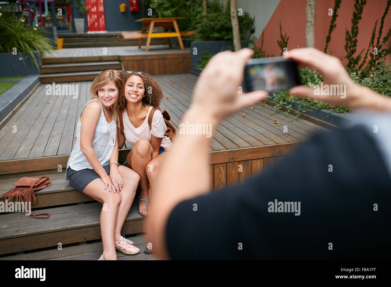 Due giovani donna seduta insieme che posano per una foto essendo tenuto da un loro amico. L'uomo prendendo fotografie dei suoi amici con mob Foto Stock