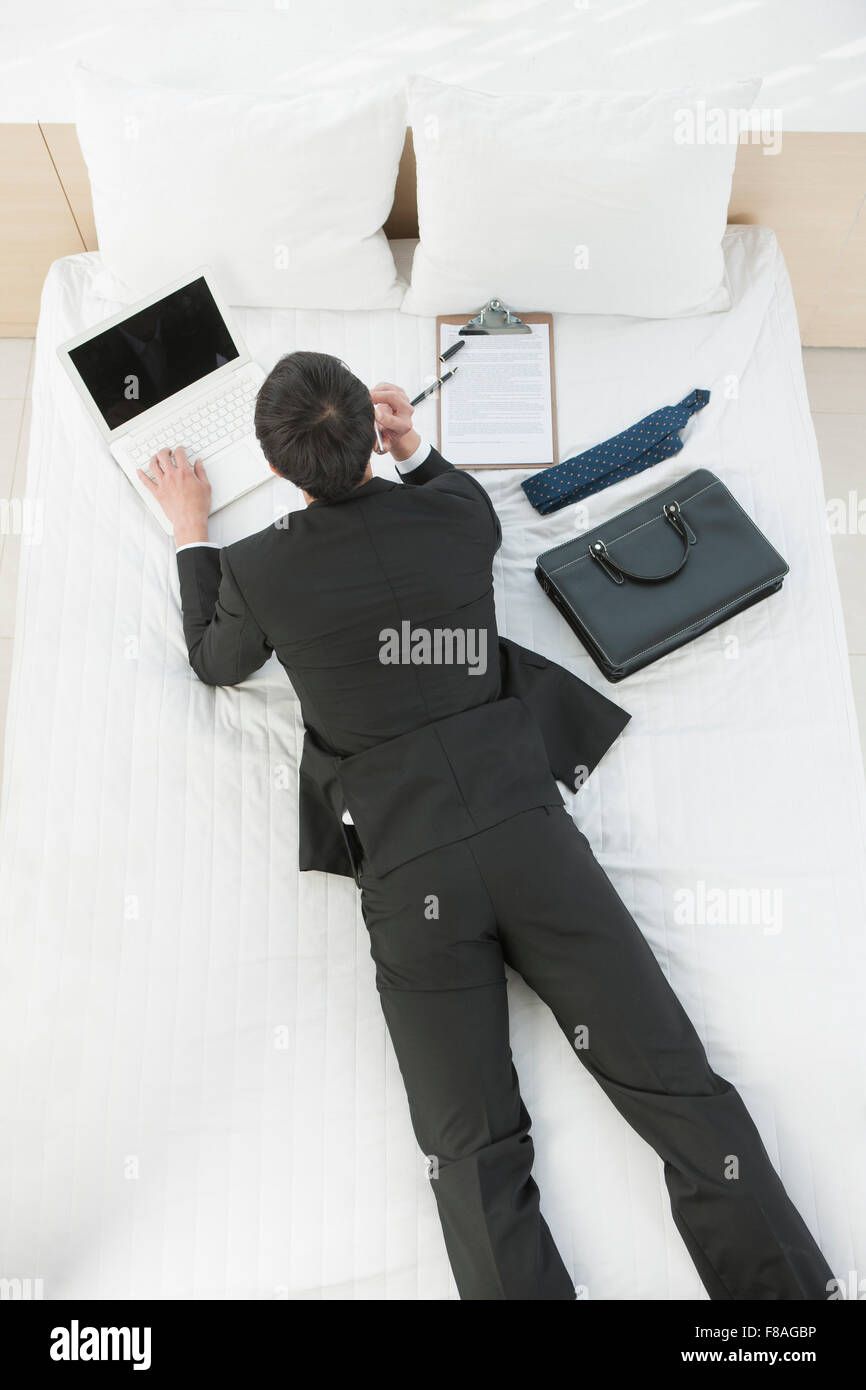 Uomo in tuta sdraiato a faccia in giù sul letto e di lavoro sul computer portatile Foto Stock