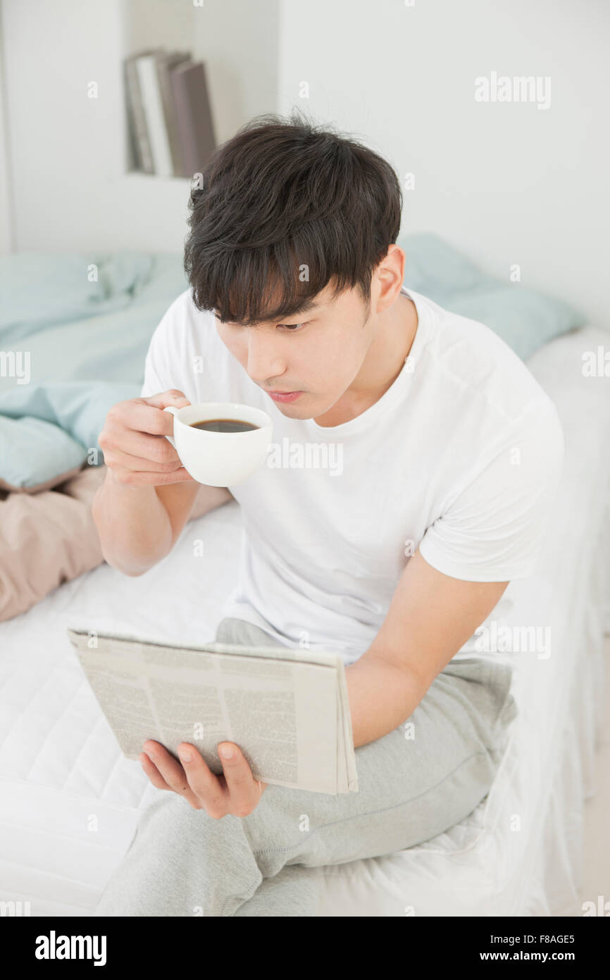 Elevato angolo di uomo seduto sul letto di bere una tazza di caffè e un giornale di lettura Foto Stock