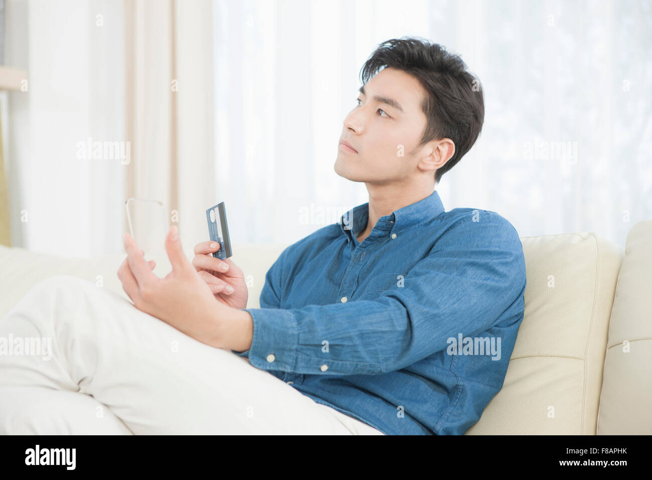 Vista laterale del giovane uomo in possesso di una carta di credito e un smartphone trasparente cercando sul divano Foto Stock