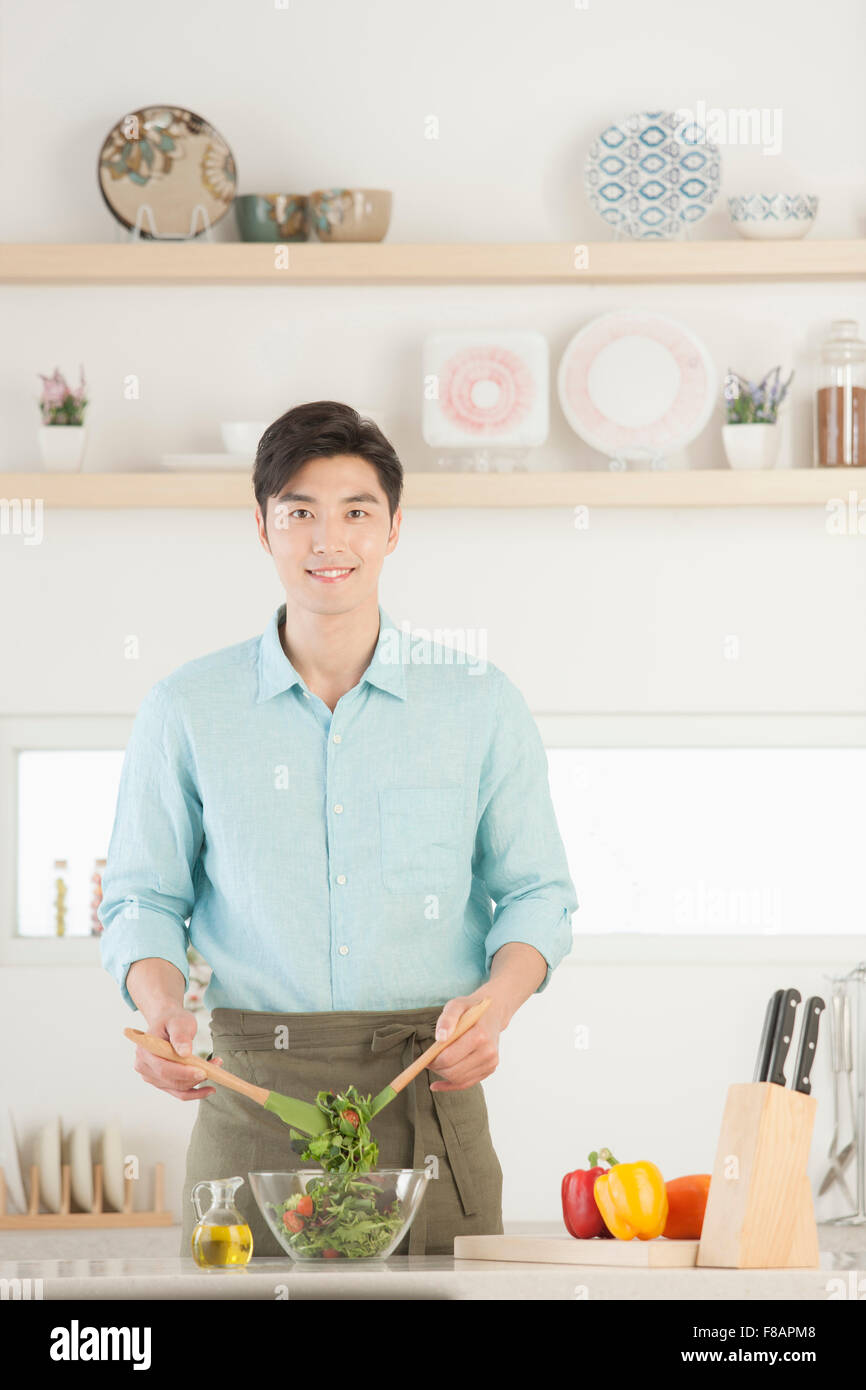 Sorridente giovane uomo in grembiule insalata di miscelazione con lo sguardo fisso anteriore in cucina Foto Stock