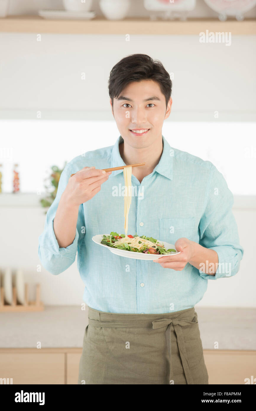 Sorridente giovane nel grembiule di prelevare la pasta con bacchette fissando davanti in cucina Foto Stock