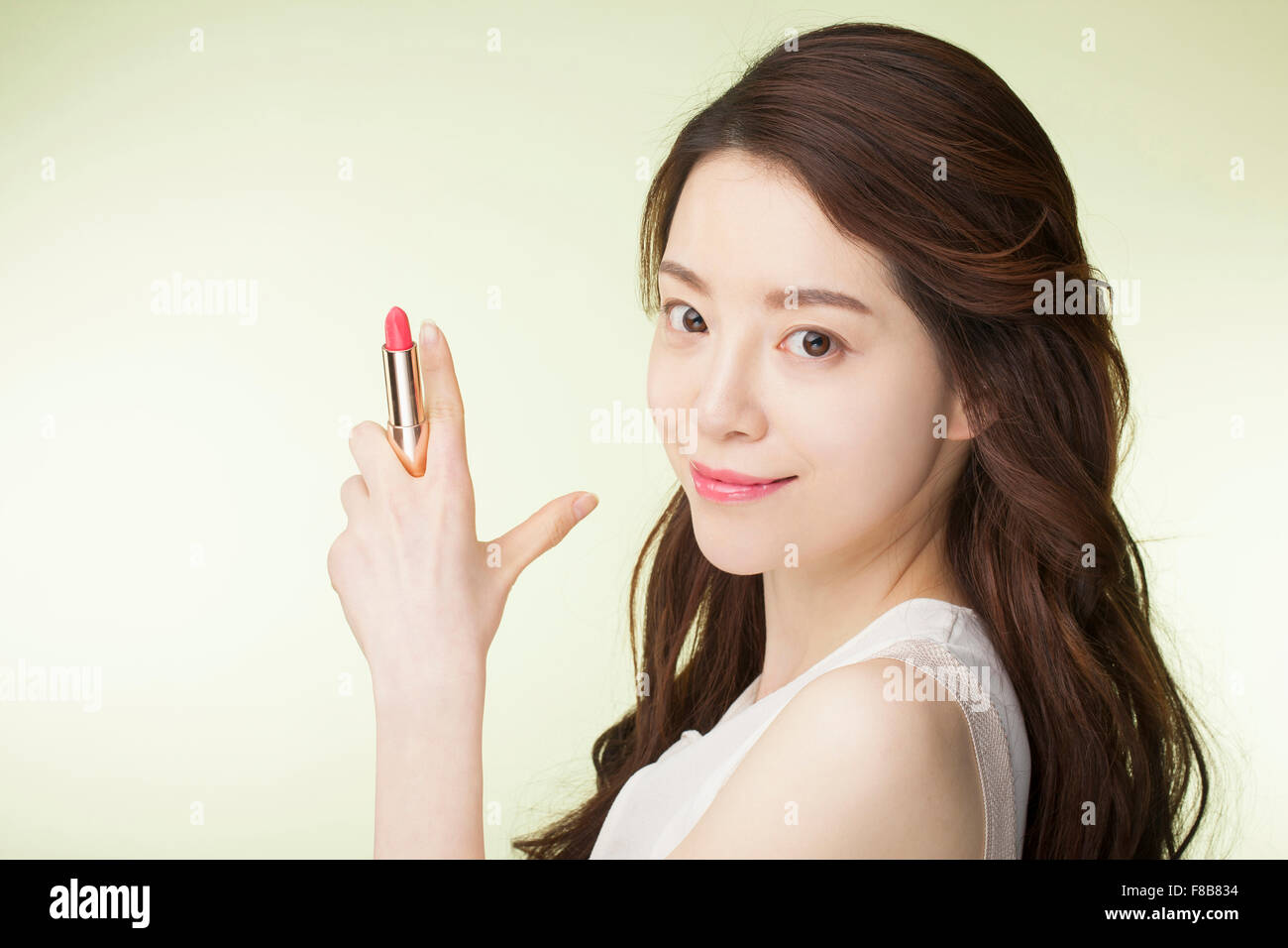 Il coreano donna con lunghi capelli ondulati in bianco camicia senza maniche tenendo un rossetto rosa con la mano il gesto e sorridente Foto Stock