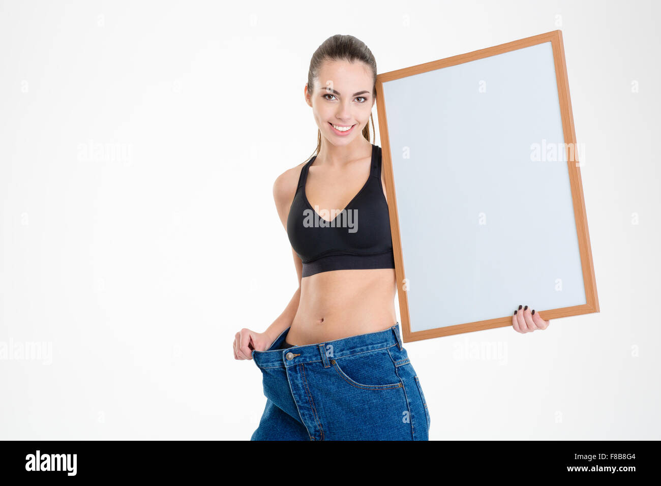 Sorridente carina ragazza fitness in vecchi jeans è diventato troppo grande azienda blank board isolate su sfondo bianco Foto Stock