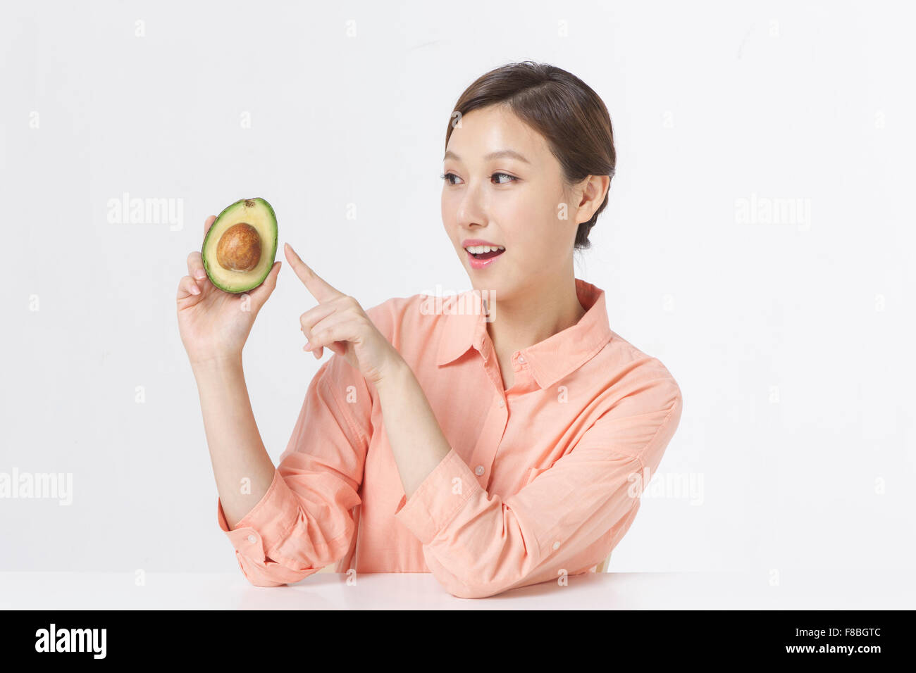 Donna adulta in maglietta arancione seduto a tavola tenendo metà di puntamento di avocado con il dito Foto Stock