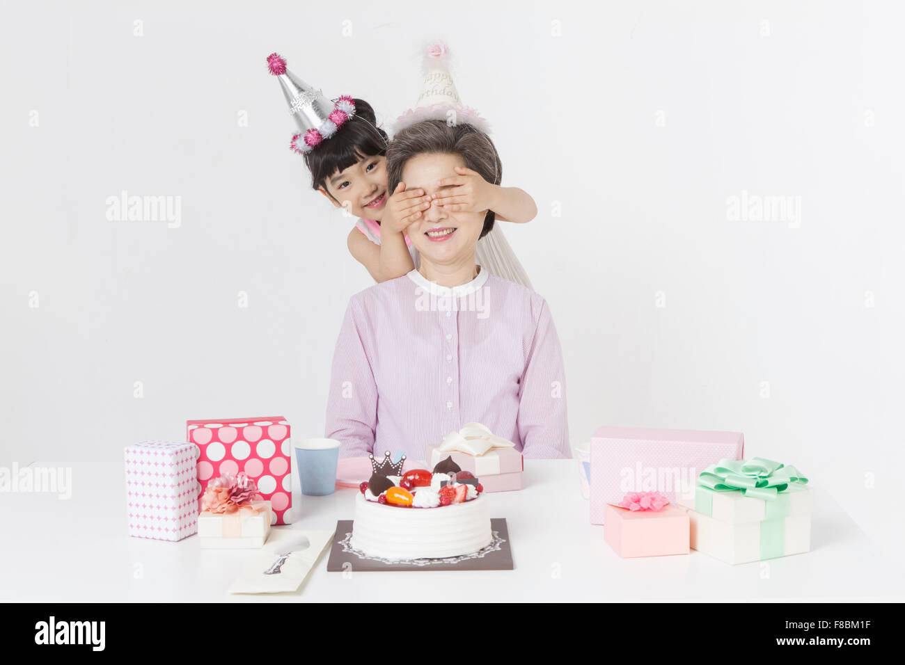 La nipote che copre la sua nonna gli occhi per festeggiare il compleanno sia in parte cappelli e confezioni regalo e una torta immessi sul Foto Stock