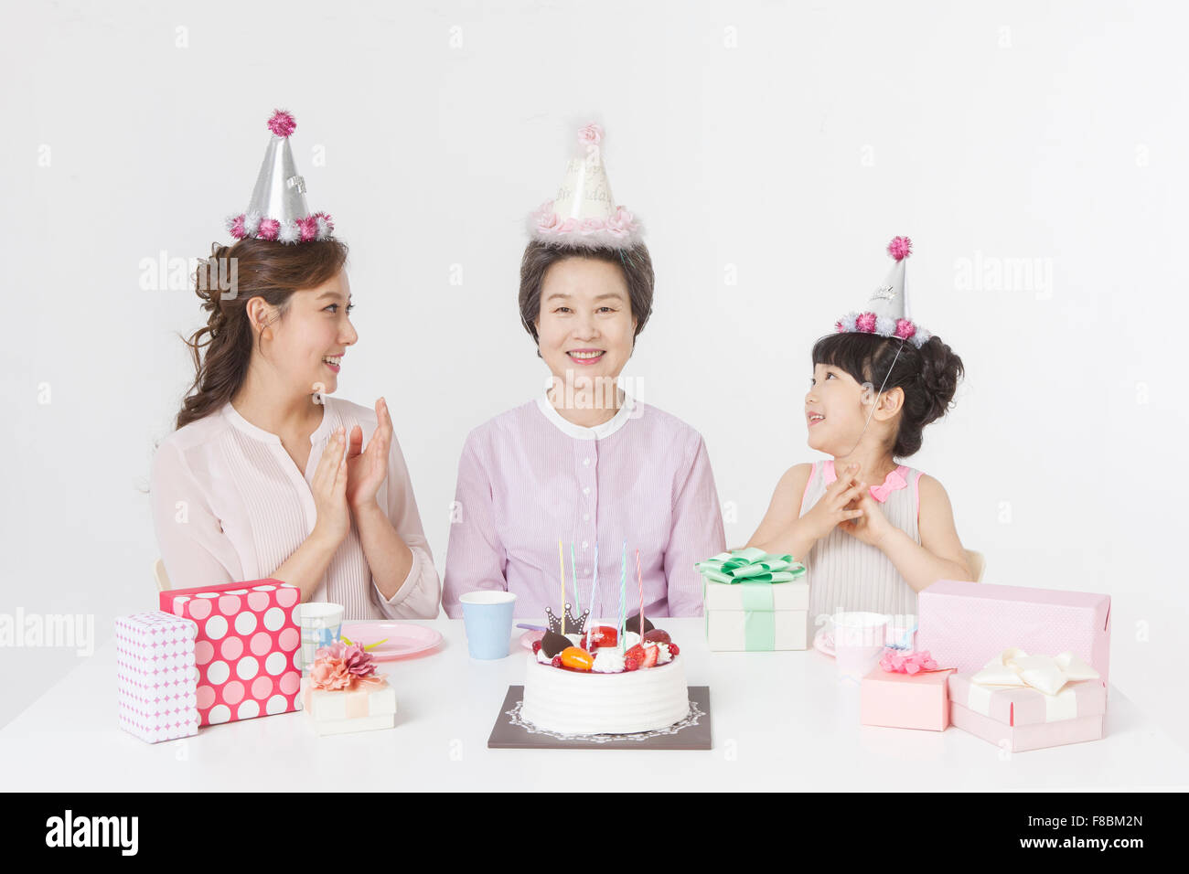 Giovane ragazza e sua madre nonna festeggia il compleanno tutti in party hat e seduti a tavola con una torta e confezioni regalo Foto Stock