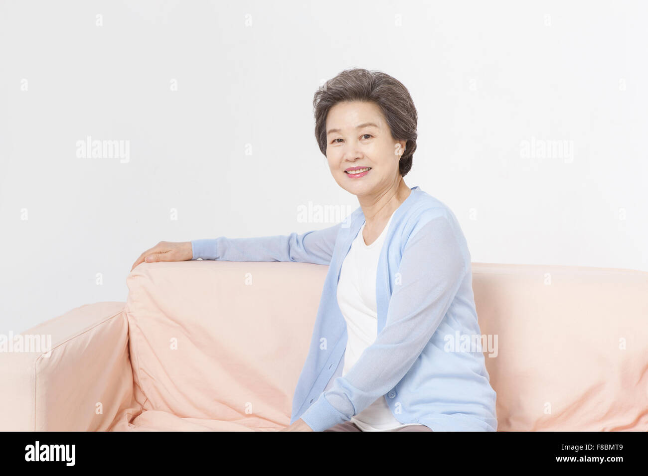 Senior donna con capelli corti in blu cardigan seduto su un divano e con lo sguardo in avanti con un sorriso Foto Stock