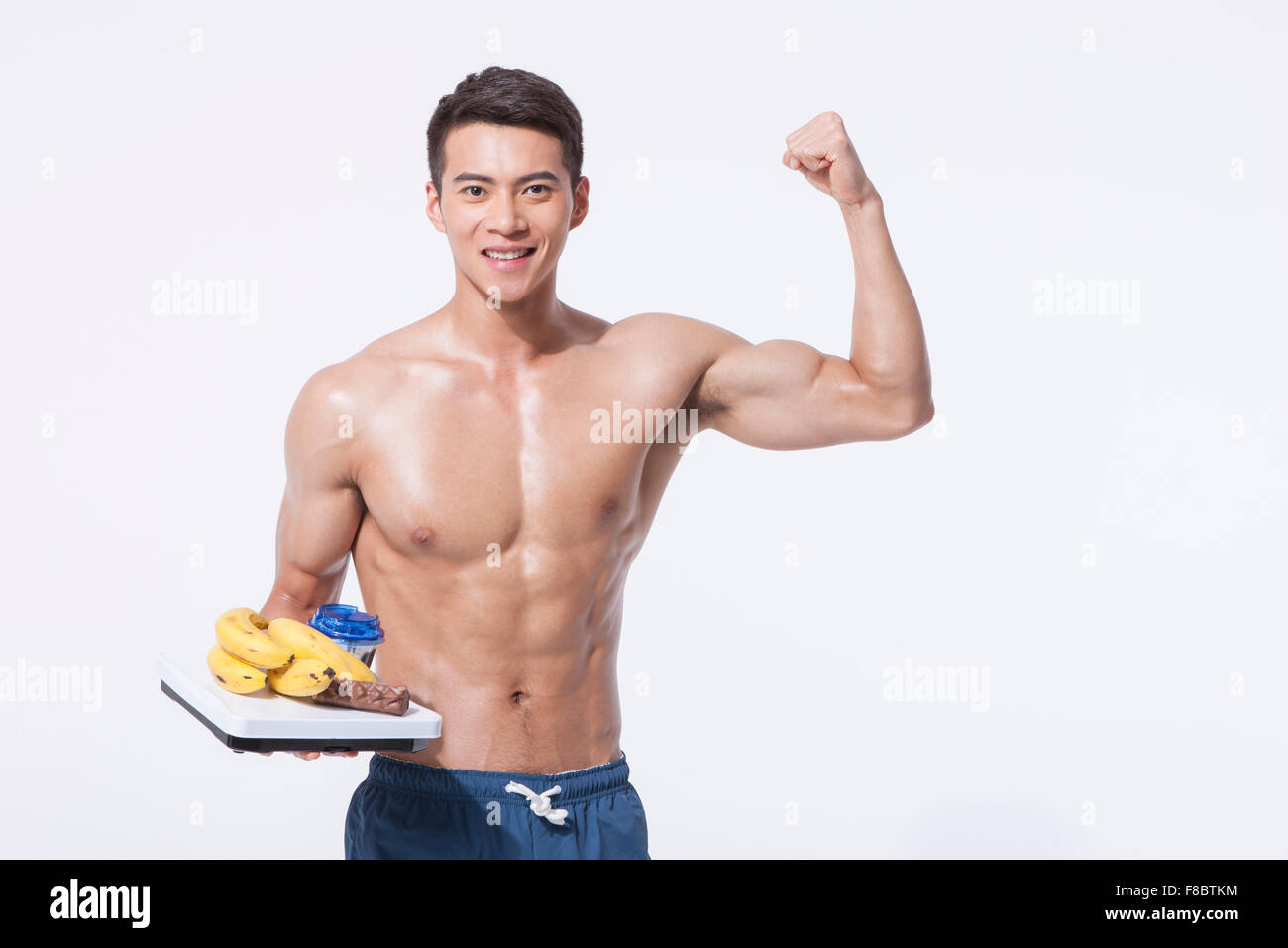 Uomo muscolare in pantaloni blu tenendo una scala con le banane e altri alimenti della dieta con una mano e porre l'altra mano mostra Foto Stock