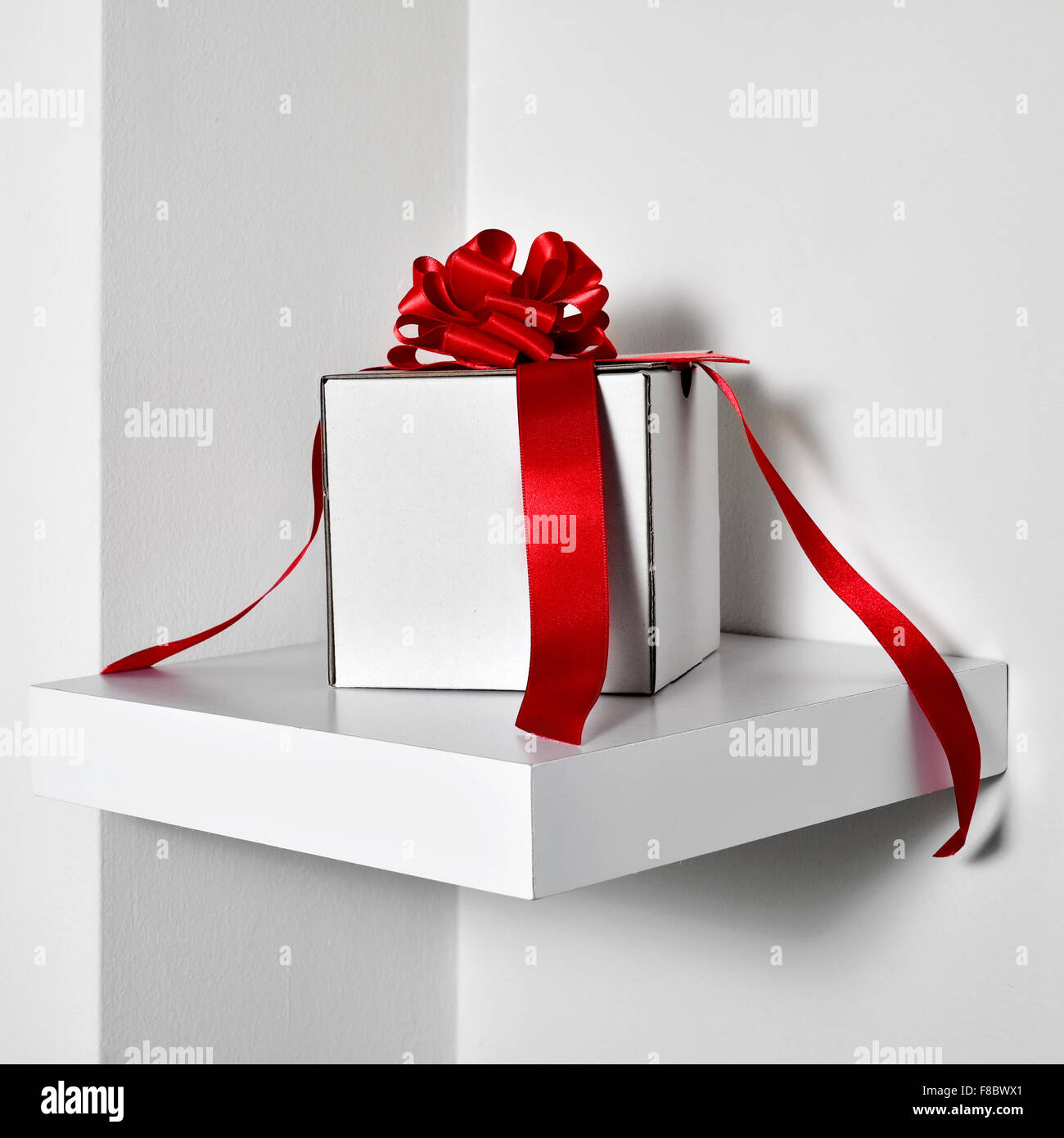 Primo piano di una scatola regalo bianca con un raso rosso nastro su un ripiano di bianco in un muro bianco Foto Stock