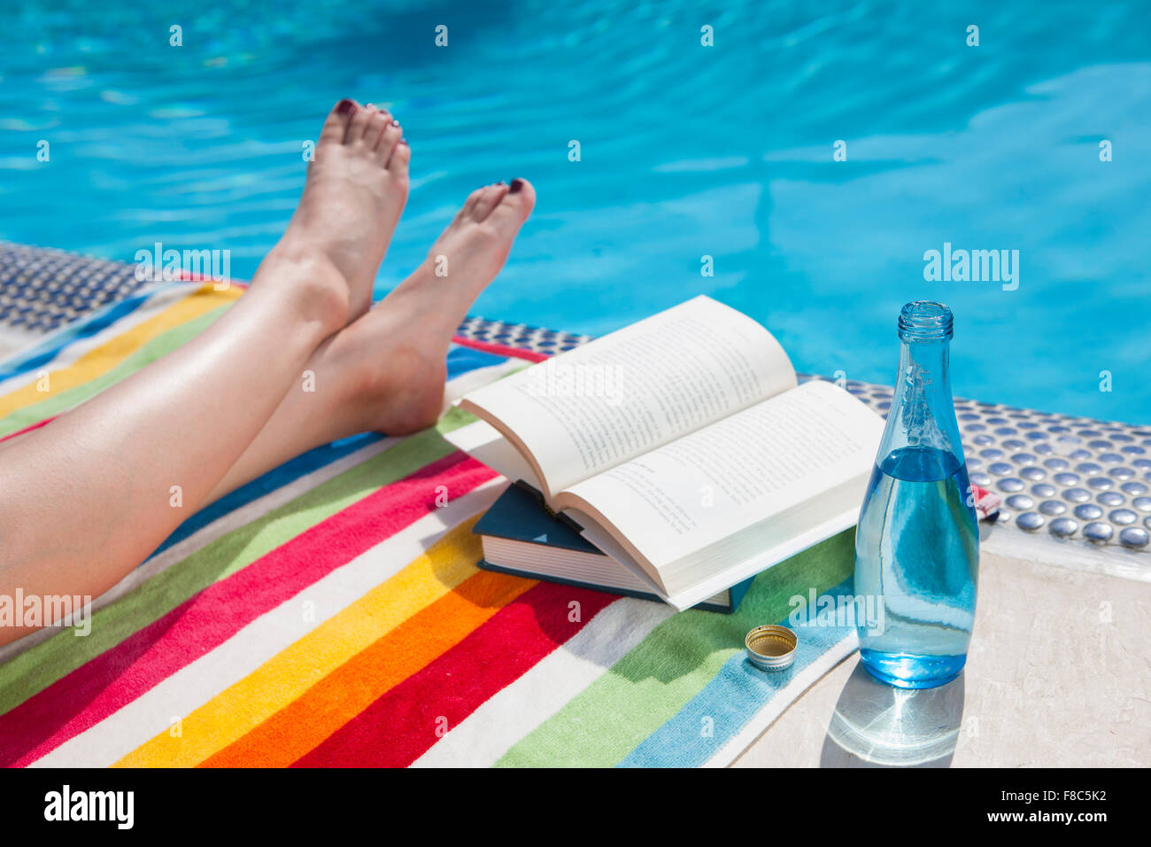 Gambe incrociate, libri e una bottiglia di bevanda con lo sfondo della piscina Foto Stock