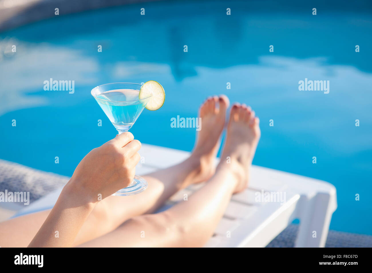 Bicchiere da cocktail con fetta di lime detenute da una sola persona che mostra una mano e le gambe con lo sfondo della piscina Foto Stock