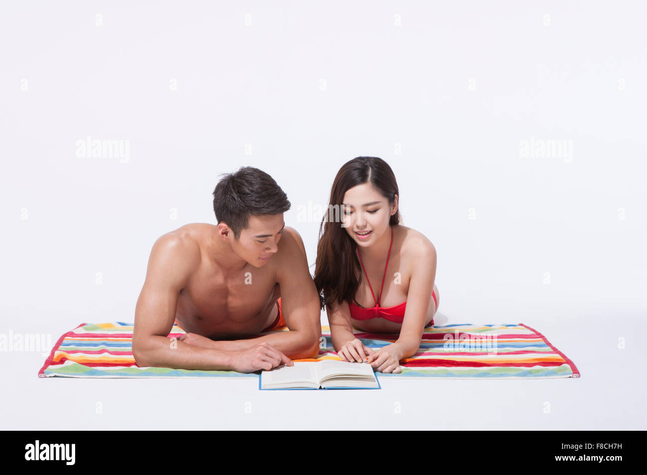 Matura in spiaggia indossare giacente sulla parte anteriore su una colorata asciugamano e guardando insieme un libro Foto Stock