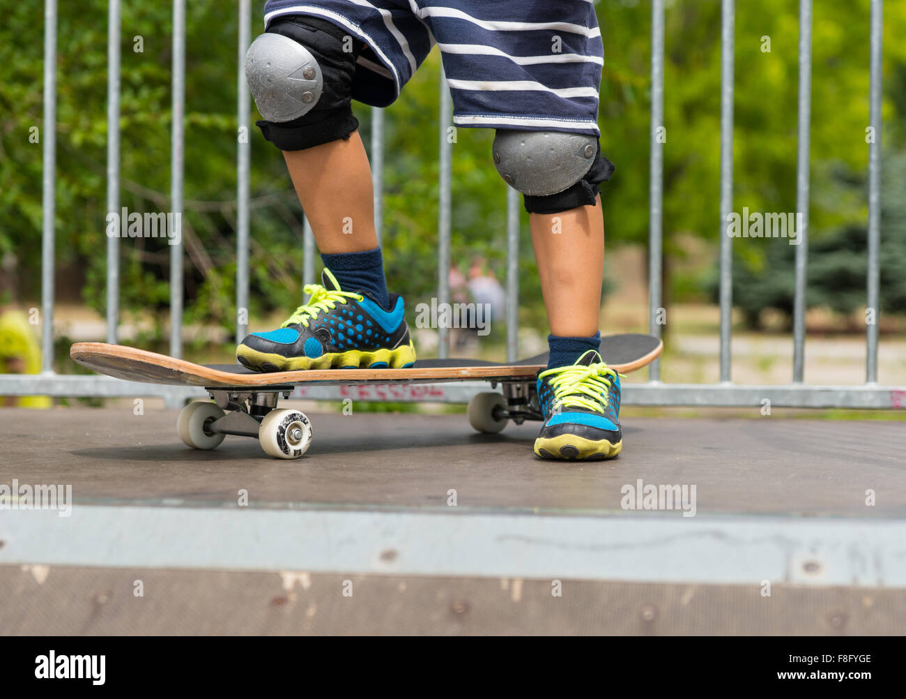 Chiusura del giovane ragazzo che indossa scarpe da ginnastica e ginocchiere  e pantaloncini a righe con un piede su skateboard alla sommità della rampa  in Skate Park sulla soleggiata giornata estiva Foto