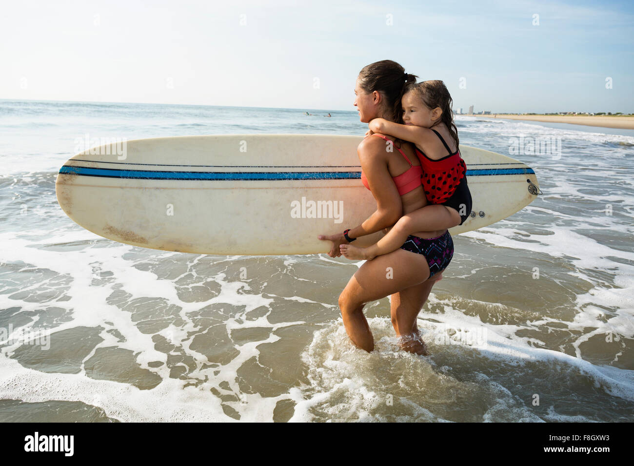 La madre che porta la figlia e la tavola da surf in spiaggia Foto Stock