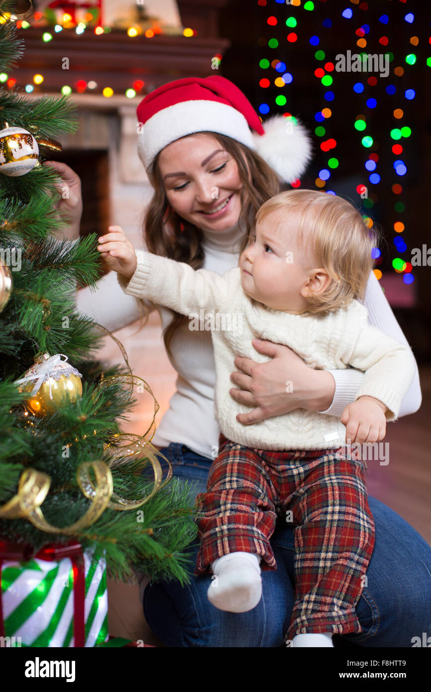 Madre in rosso santa hat guarda al suo piccolo figlio toddler e sorridente nei pressi di alberi di Natale Foto Stock