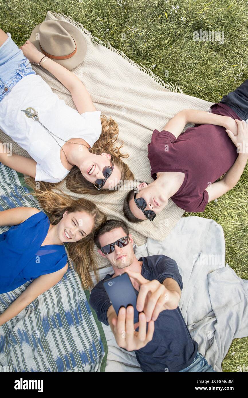 Un gruppo di giovani adulti giacente su coperte da picnic, tenendo autoritratto utilizza lo smartphone Foto Stock