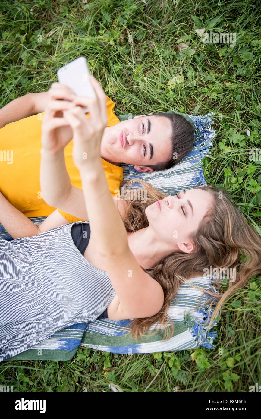 Coppia giovane giacente su erba in campo, tenendo autoritratto utilizza lo smartphone Foto Stock