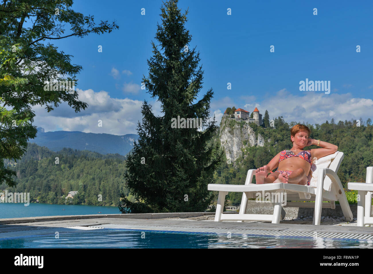 Conciato al bianco di mezza età della donna a prendere il sole su una sedia a sdraio vicino alla piscina esterna. Lago di Bled e del castello in background. Foto Stock