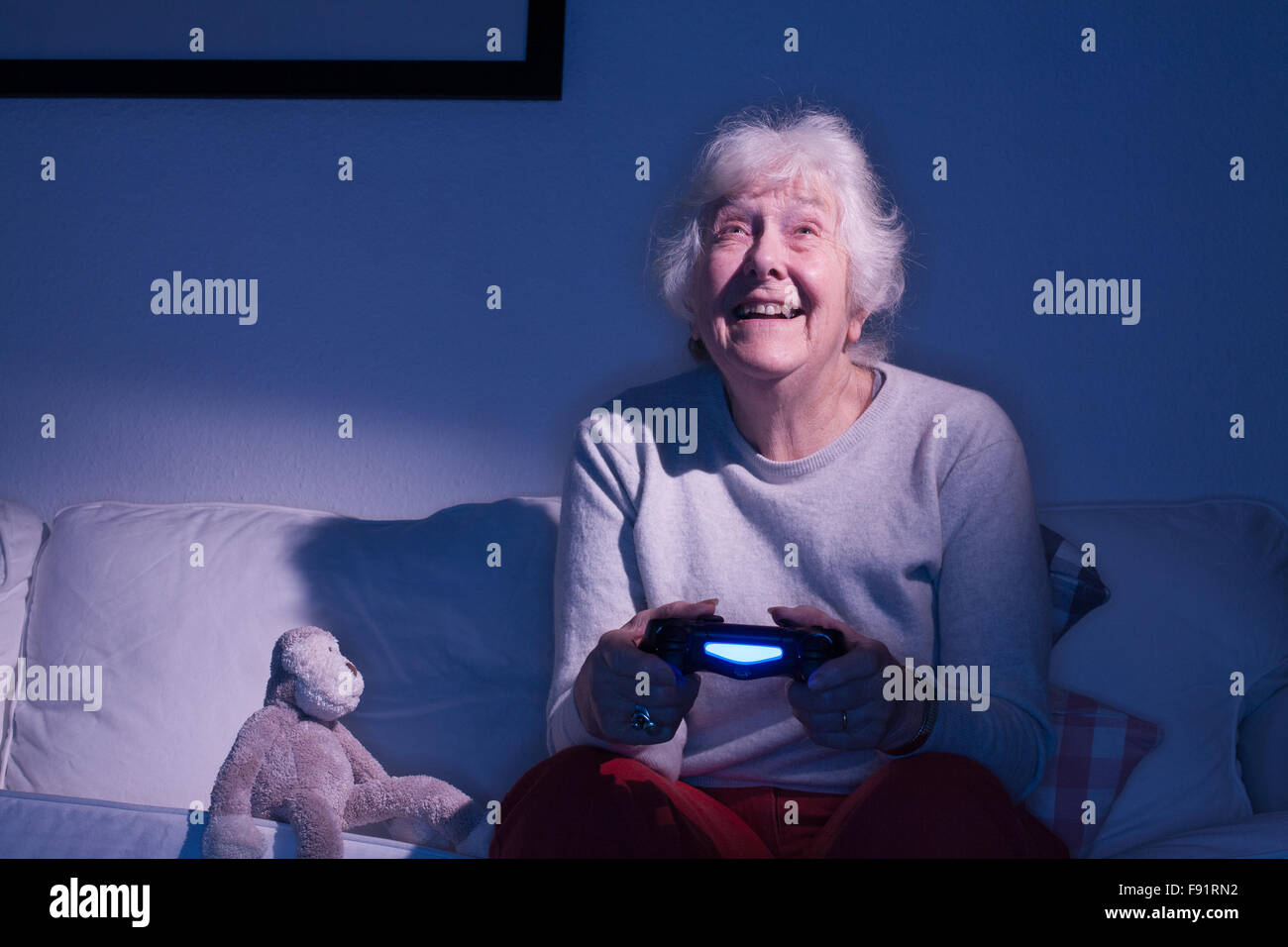 Anziana signora seduta su un divano a giocare su PLAYSTATION 4 il prossimo ad un giocattolo imbottito scimmia. Foto Stock