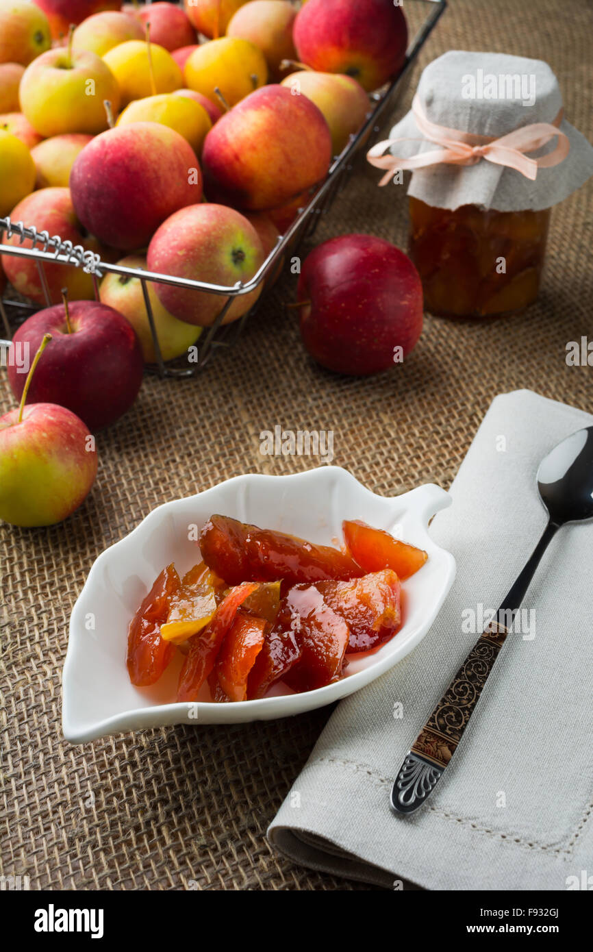 In casa fette di mela marmellata su bianco piattino, cucchiaio, un vasetto di marmellata di arance, biancheria igienico e mele in filo metallico cesto su una tela cov Foto Stock